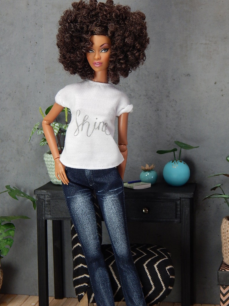 May include: A doll with brown curly hair wearing a white t-shirt with the word "shine" printed on it and blue jeans.