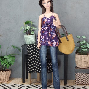 May include: A doll wearing a purple floral print top, blue jeans, and black heels. The doll is holding a brown leather tote bag. The doll is standing on a black and white patterned rug in front of a gray wall.