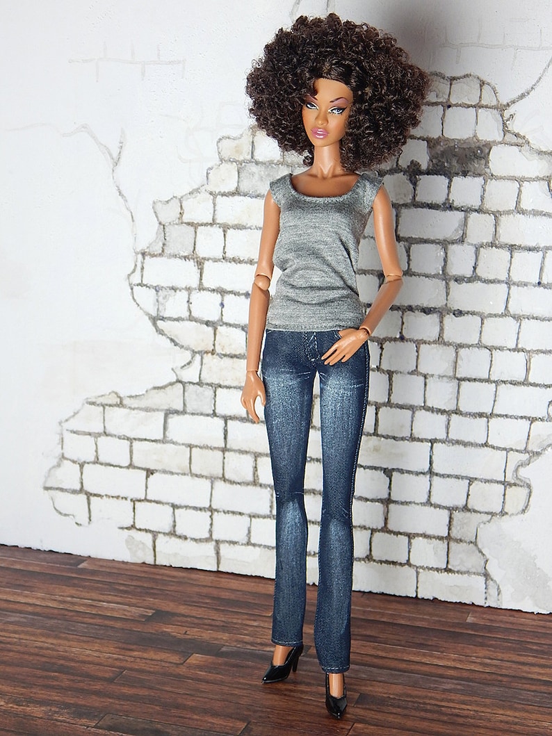May include: A fashion doll with brown curly hair wearing a gray tank top and blue jeans. The doll is standing in front of a white brick wall.