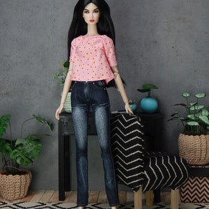 May include: A doll wearing a pink and orange polka dot top and blue jeans. The doll has long black hair and is standing in front of a chair with a black and white chevron pattern.