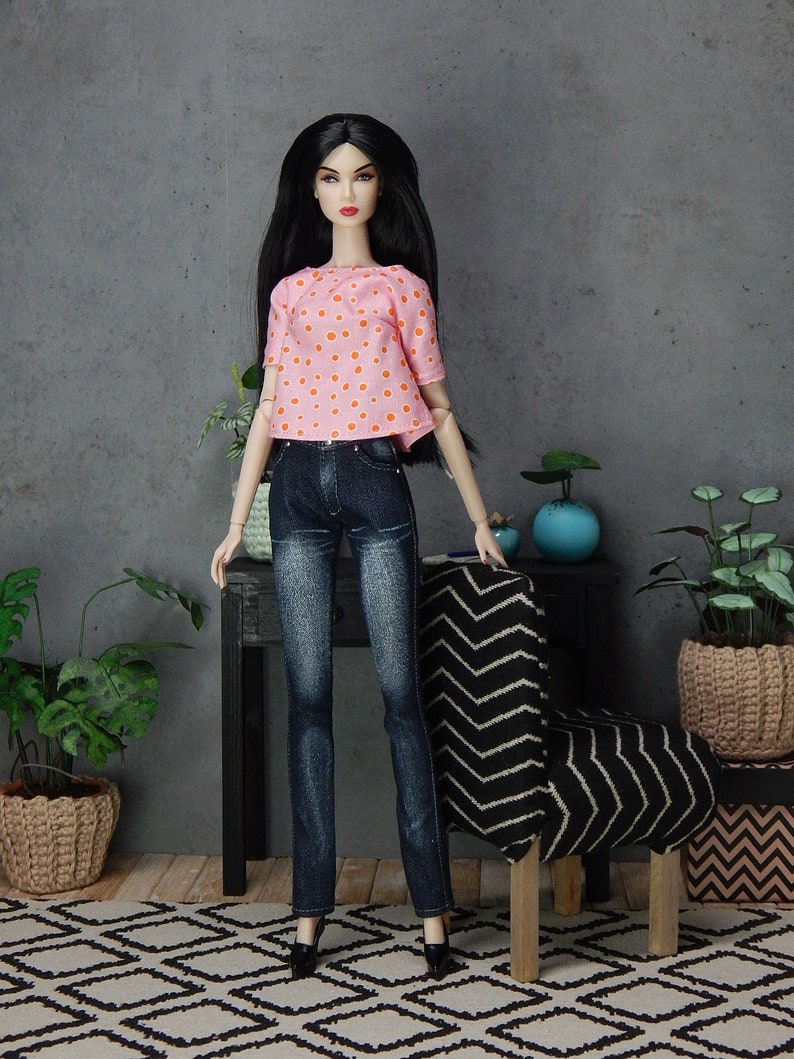 May include: A doll wearing a pink and orange polka dot top and blue jeans. The doll has long black hair and is standing in front of a chair with a black and white chevron pattern.