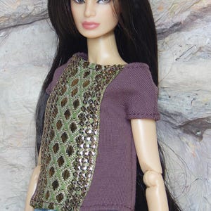 May include: A fashion doll with long dark hair wearing a short-sleeved purple top featuring a central panel of green and metallic bronze geometric embroidery with sequins, paired with a light blue skirt decorated with a small white floral pattern.