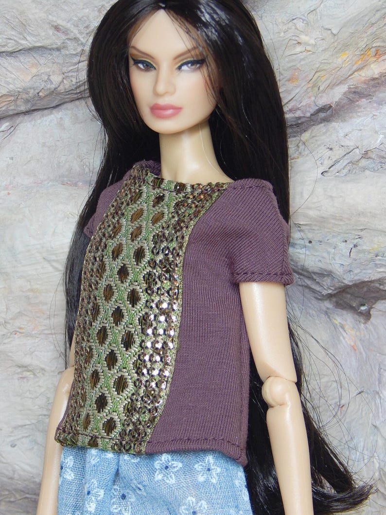 May include: A fashion doll with long dark hair wearing a short-sleeved purple top featuring a central panel of green and metallic bronze geometric embroidery with sequins, paired with a light blue skirt decorated with a small white floral pattern.