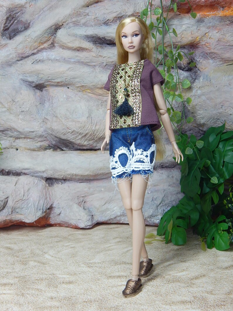 May include: A fashion doll with long blonde hair wearing a brown short-sleeved top with a gold sequined front panel and a dark blue denim skirt featuring a white lace trim at the hem. The doll is wearing metallic bronze slip-on shoes.