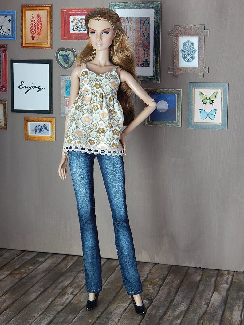 May include: A doll wearing a white and brown floral print tank top with a lace trim, blue jeans, and black heels. The doll has long brown hair and is standing in front of a wall with framed pictures.