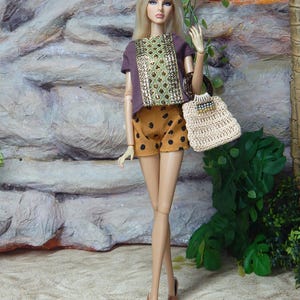 May include: A fashion doll with long blonde hair wearing a purple short-sleeved top with a central gold sequined panel, mustard yellow shorts with black polka dots, and tan high-heeled shoes. The doll carries a beige crocheted handbag with a dark handle.