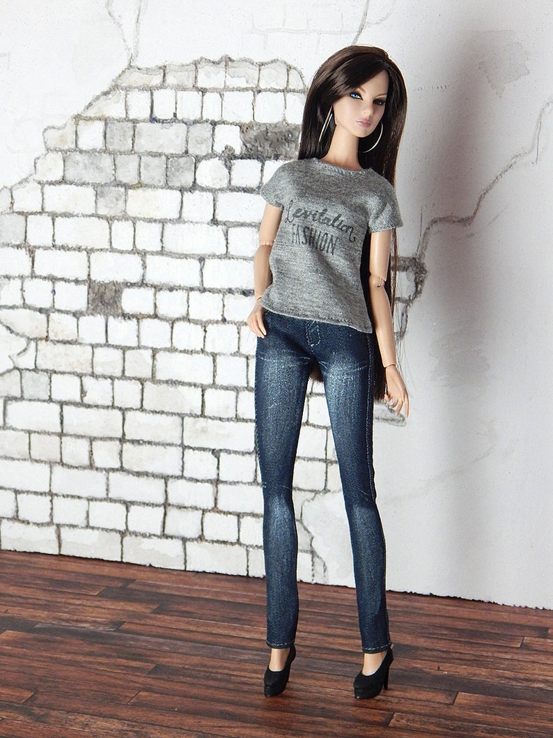 May include: A doll wearing a grey t-shirt with the text "Celebrating Fashion" and blue jeans. The doll has long brown hair and is wearing black heels.