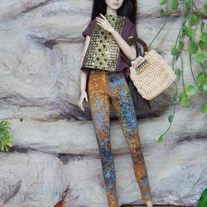 May include: A fashion doll with long dark hair wearing a brown short-sleeved top with a gold sequined front panel, multi-colored speckled leggings in shades of blue and orange, and black strappy heels, carrying a beige woven handbag with a dark handle.