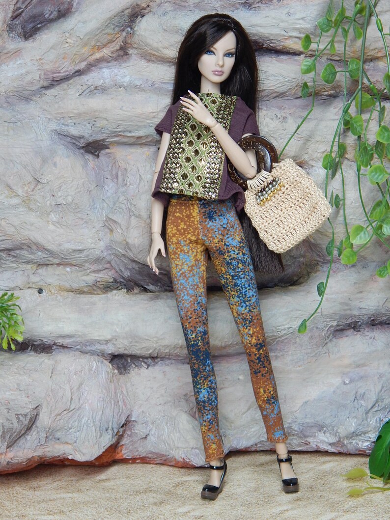 May include: A fashion doll with long dark hair wearing a brown short-sleeved top with a gold sequined front panel, multi-colored speckled leggings in shades of blue and orange, and black strappy heels, carrying a beige woven handbag with a dark handle.