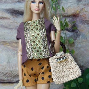 May include: A fashion doll with long blonde hair wearing a purple short-sleeved top with a central gold and green textured panel, paired with mustard yellow shorts featuring black polka dots and a beige crocheted handbag with a dark circular handle.