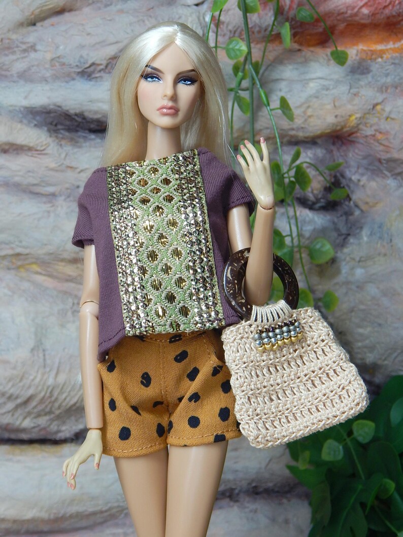 May include: A fashion doll with long blonde hair wearing a purple short-sleeved top with a central gold and green textured panel, paired with mustard yellow shorts featuring black polka dots and a beige crocheted handbag with a dark circular handle.