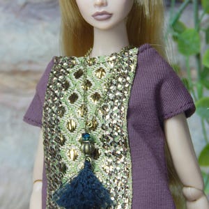 May include: A doll with long blonde hair wearing a short-sleeved plum-colored top featuring a central panel of metallic gold and light green woven fabric. A long necklace with a dark blue tassel and a small blue bead hangs over the chest.