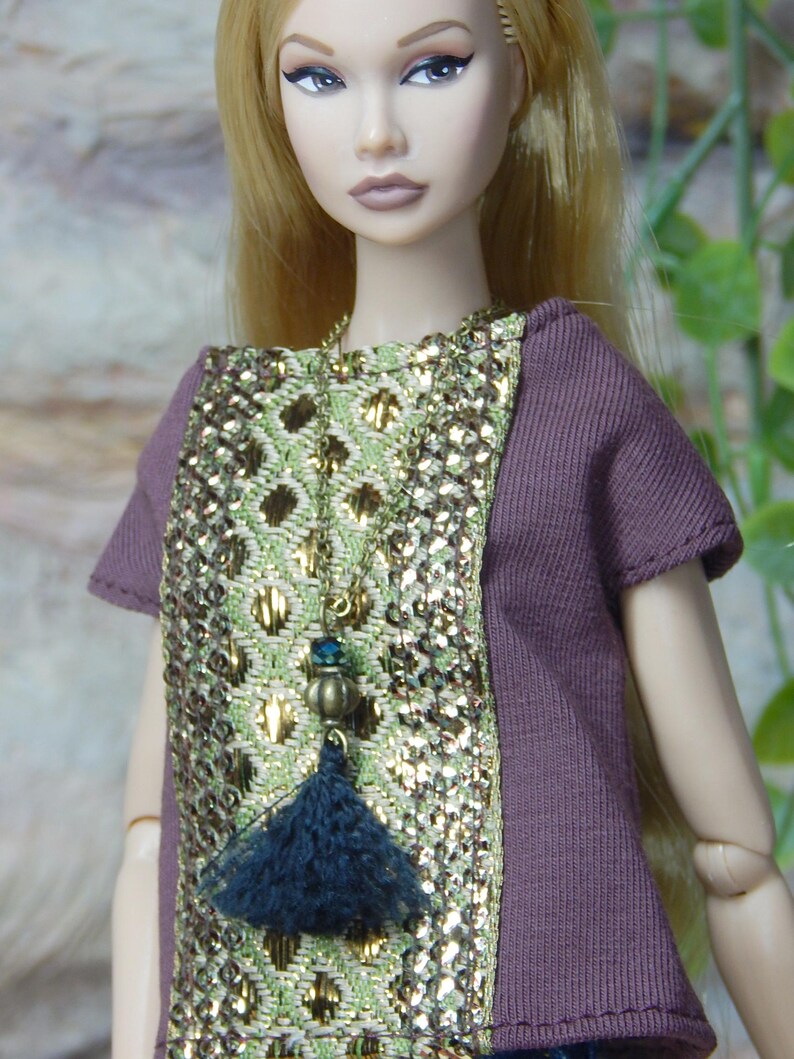 May include: A doll with long blonde hair wearing a short-sleeved plum-colored top featuring a central panel of metallic gold and light green woven fabric. A long necklace with a dark blue tassel and a small blue bead hangs over the chest.