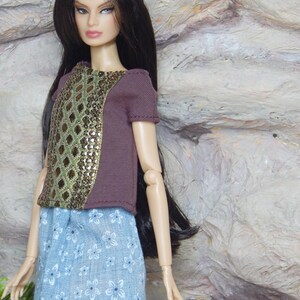 May include: A fashion doll with long dark hair wearing a short-sleeved brown top featuring a central panel with a gold and green diamond pattern. The doll is also wearing a light blue skirt with a white floral print and has articulated joints.