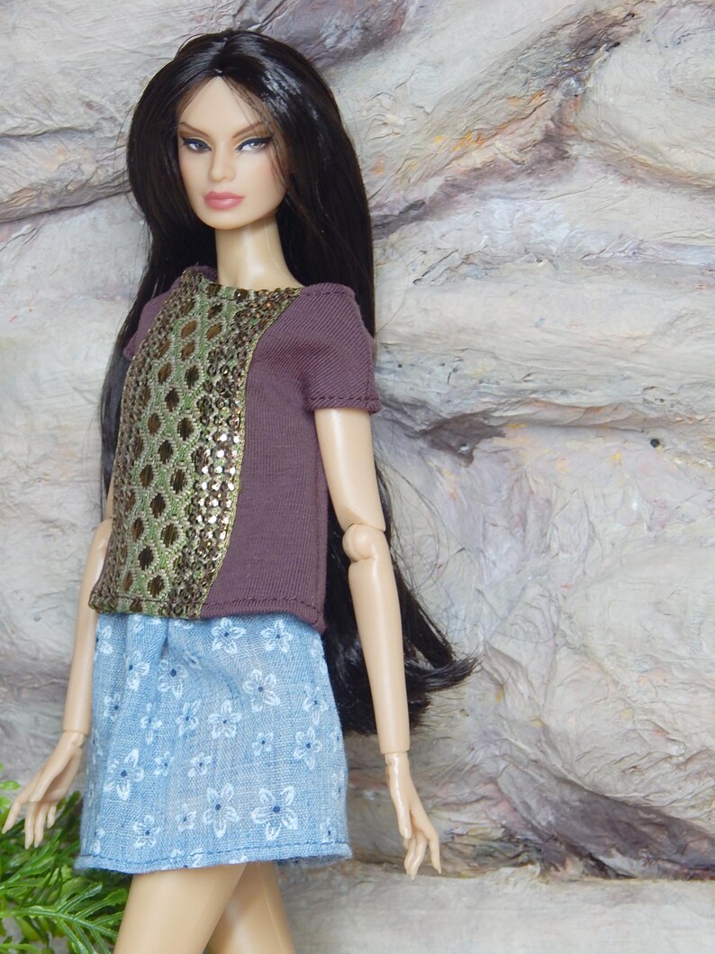 May include: A fashion doll with long dark hair wearing a short-sleeved brown top featuring a central panel with a gold and green diamond pattern. The doll is also wearing a light blue skirt with a white floral print and has articulated joints.