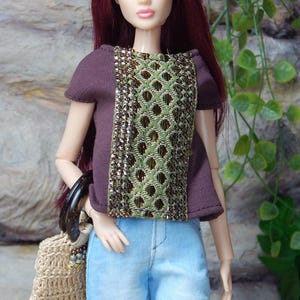 May include: A doll with long burgundy hair wearing a short-sleeved dark brown top featuring a central panel of green and gold geometric embroidery, paired with light blue denim pants and carrying a small beige crocheted handbag with dark handles.