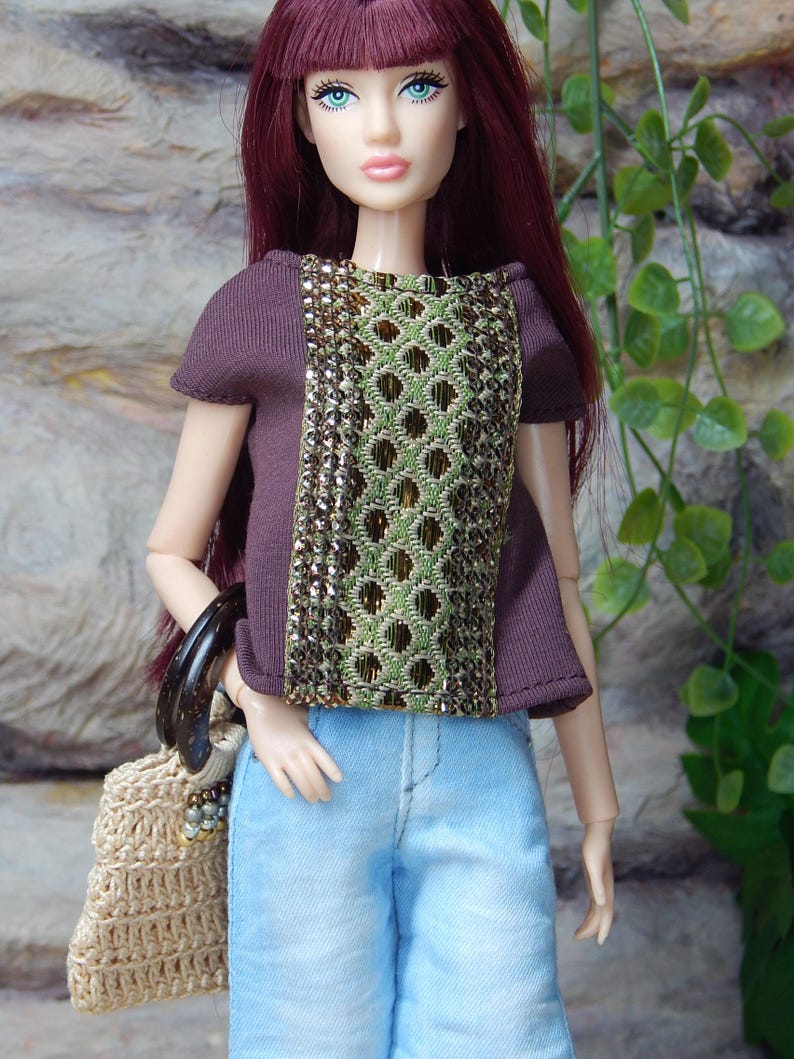 May include: A doll with long burgundy hair wearing a short-sleeved dark brown top featuring a central panel of green and gold geometric embroidery, paired with light blue denim pants and carrying a small beige crocheted handbag with dark handles.