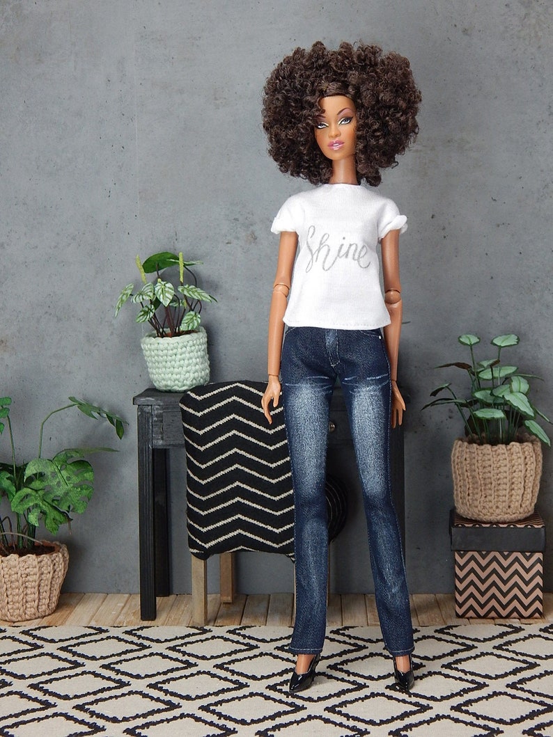 May include: A doll with brown curly hair wearing a white t-shirt with the word "shine" on it, blue jeans, and black shoes. The doll is standing on a black and white patterned rug in front of a black table with a black and white patterned throw blanket.