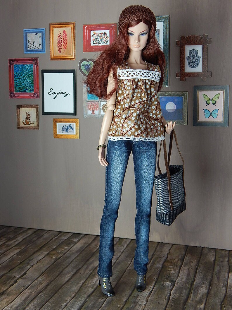 May include: A doll wearing a brown and white patterned top with a white lace trim, blue jeans, a brown knitted hat, and black shoes. The doll is holding a brown and grey tote bag. The doll is standing in front of a wall with framed pictures.