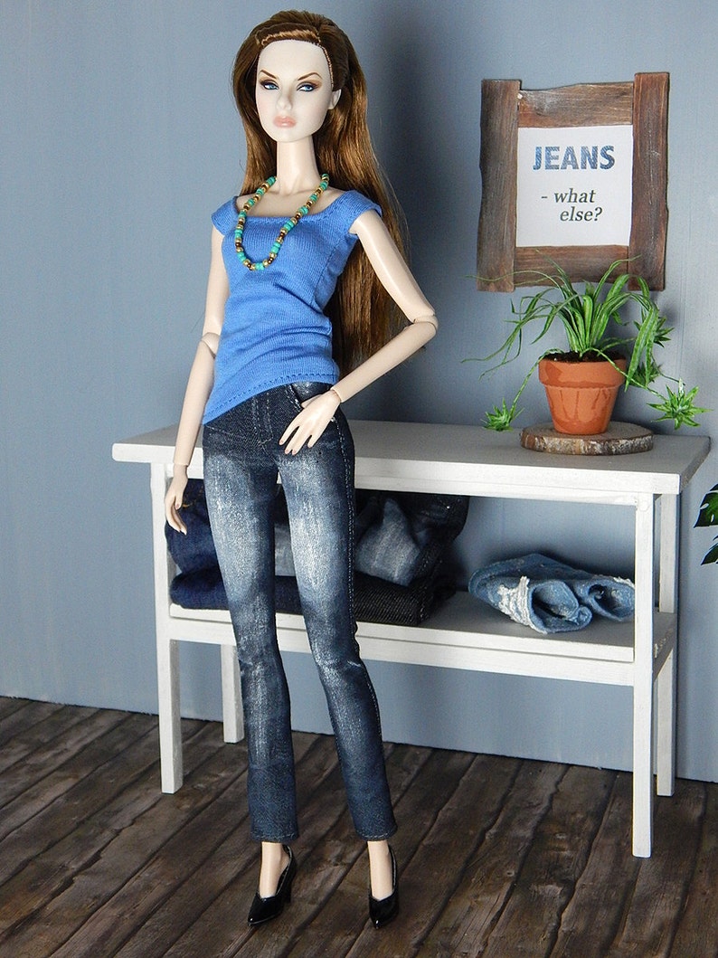 May include: A doll wearing a blue tank top with a turquoise and gold beaded necklace and blue jeans. The doll is standing in front of a white shelf with a sign that reads "JEANS - what else?"