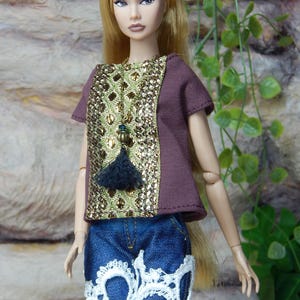 May include: A fashion doll with long blonde hair wearing a short-sleeved brown top featuring a central panel of gold metallic sequins and a dark tassel pendant. The doll wears blue denim shorts with frayed hems and white lace trim at the bottom.