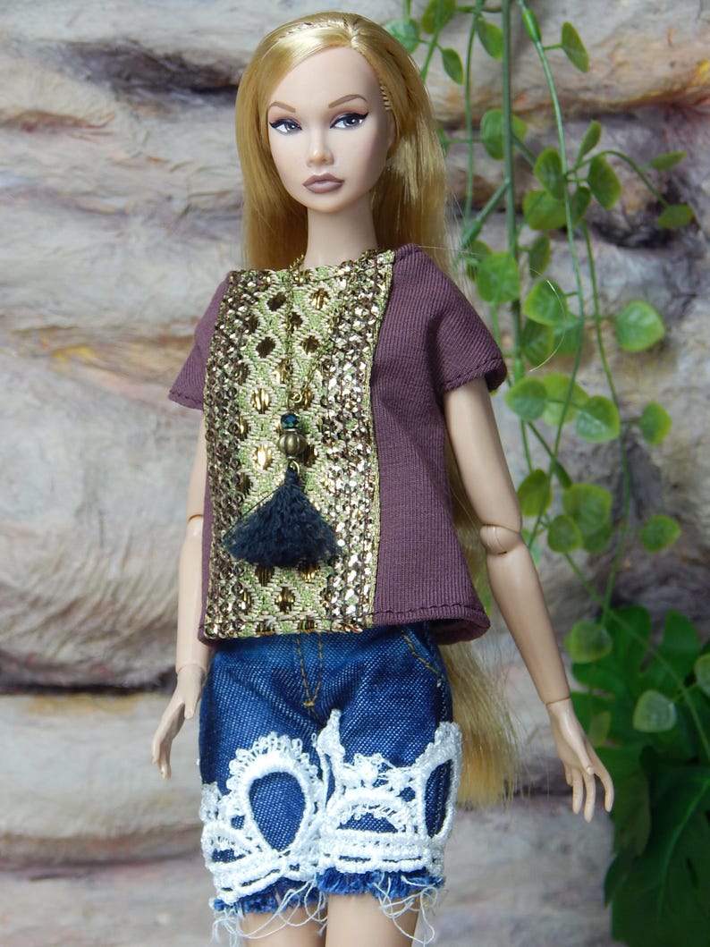 May include: A fashion doll with long blonde hair wearing a short-sleeved brown top featuring a central panel of gold metallic sequins and a dark tassel pendant. The doll wears blue denim shorts with frayed hems and white lace trim at the bottom.