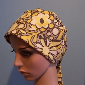May include: A yellow and brown floral print fabric head covering with a tie. The fabric has a pattern of large and small flowers.