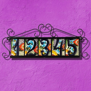May include: Decorative house number sign featuring the numbers 1, 2, 3, 4, and 5. Each number is black and set against a colorful floral tile background with red, blue, and yellow flowers. The sign is framed by a black wrought iron scroll design.