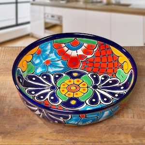 May include: A vibrant, hand-painted ceramic bowl with a blue rim and intricate floral designs in red, blue, green, and yellow. The bowl sits on a wooden surface, showcasing its colorful, decorative pattern.