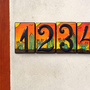 May include: Four square ceramic house number tiles. Each tile features a black number against a desert sunset backdrop with cacti. The numbers are 1, 2, 3, and 4. The tiles are mounted on a light-colored wall.