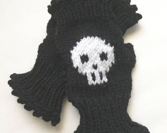 May include: Black knitted fingerless gloves with a white skull design. The gloves have a scalloped edge and are made of a thick, textured yarn. The skull is centered on the back of the hand.