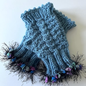 May include: A pair of light blue knitted fingerless gloves. The gloves have a textured pattern and are trimmed with black fringe and colorful accents. The cuffs have a scalloped edge.