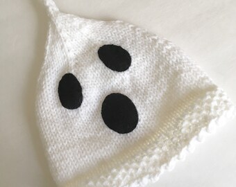 May include: A white knit hat with three large black oval shapes, resembling eyes, is displayed on a white surface. The hat has a pointed top with a knotted detail. The bottom edge has a scalloped trim, creating a ghost-like appearance.