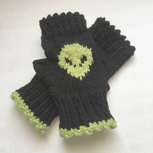 May include: Black fingerless gloves with a lime green skull design. The gloves are knitted with a ribbed cuff and a scalloped edge in lime green. The skull is centered on the back of the hand.