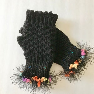 May include: A pair of dark gray knitted fingerless gloves with a textured pattern. The cuffs have a ruffled edge, and the bottom edges are trimmed with black fringe and colorful yarn accents. These gloves are designed to keep hands warm.