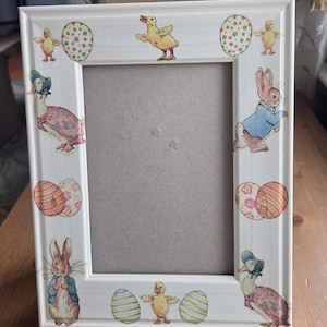 May include: White rectangular picture frame decorated with Easter-themed illustrations. The frame features images of ducks, bunnies, and decorated eggs. The frame is designed with a light wood grain finish.