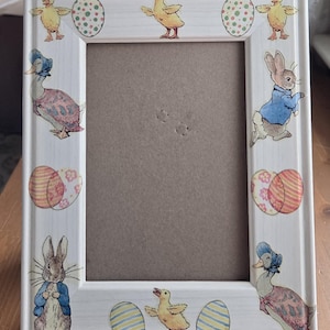 May include: White rectangular picture frame decorated with Easter-themed illustrations. The frame features images of yellow chicks, decorated eggs, a rabbit in a blue jacket, and a duck in a bonnet. The frame is designed for a standard-sized photo.