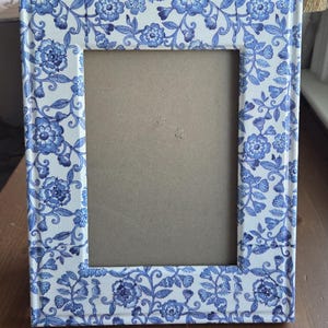 May include: A rectangular picture frame featuring a white background with a repeating blue floral pattern. The frame has a smooth, glossy finish and a centered rectangular opening with a plain tan backing visible inside the frame structure.