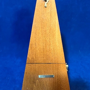 May include: A wooden metronome with a triangular shape. The metronome is made of light brown wood and has a small metal plate that reads "Seth Thomas". A key is attached to the top.