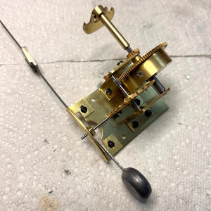 May include: A gold-toned metal clockwork mechanism with a winding key and a small, round, gray weight. The mechanism is mounted on a green metal base.