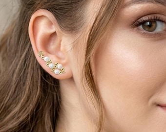 May include: Gold-toned ear climbers with white, round, pearl-like accents. The earrings curve along the ear, with a spiral design at the top. The jewelry is worn on the ear of a person with light skin and brown hair.