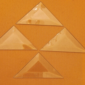 May include: Four clear, triangular glass pieces arranged on an orange fabric surface. Each triangle has a beveled edge, creating a prism effect. The glass appears to be slightly tinted, reflecting the warm background.