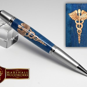May include: A silver pen with a blue and wood barrel featuring a laser engraved medical symbol. The symbol is a snake wrapped around a staff with wings. The pen is on a white background.