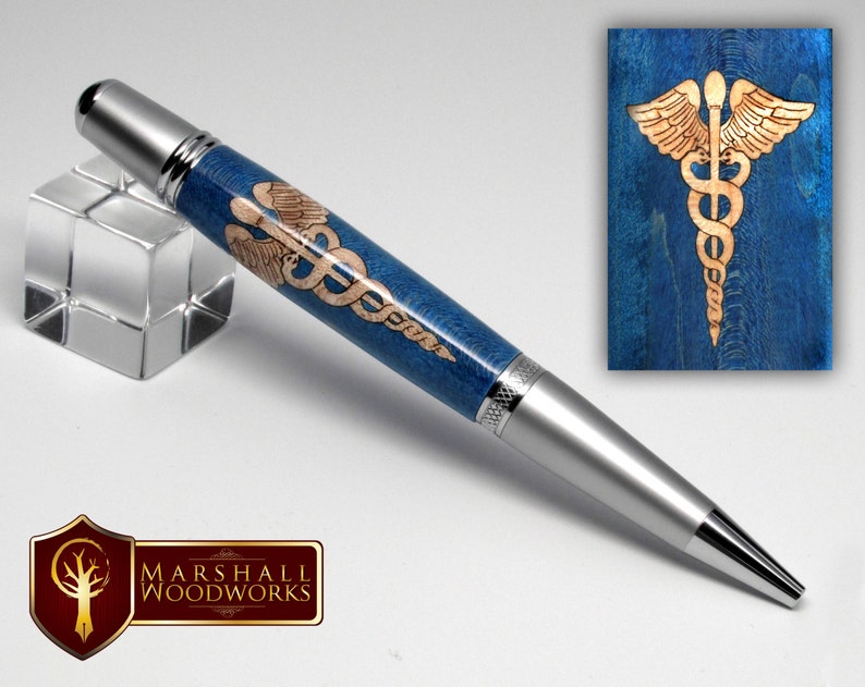 May include: A silver pen with a blue and wood barrel featuring a laser engraved medical symbol. The symbol is a snake wrapped around a staff with wings. The pen is on a white background.