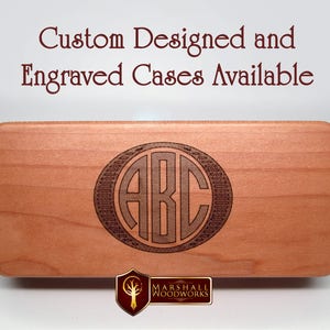 May include: A close-up of a wooden box with a custom engraved monogram of the letters "ABC" in a circle. The box is made of light wood and has a smooth finish. The engraving is dark brown and is centered on the box. The text "Custom Designed and Engraved Cases Available" is visible at the top of the image.