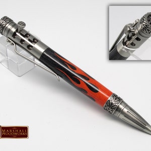 May include: A silver and black pen with a flame design on the barrel. The pen has a silver clip and a silver cap with a detailed design. The pen is on a white background.