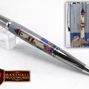 May include: A silver and wood pen featuring a lighthouse design. The pen has a blue and white background with a red and white lighthouse in the centre. The lighthouse is on a small island with birds flying around it.