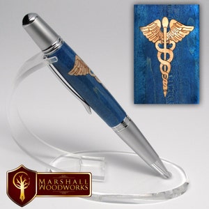 May include: A blue and silver pen with a wood inlay of the medical symbol, the caduceus. The pen is on a clear acrylic stand. The stand has a logo for Marshall Woodworks.