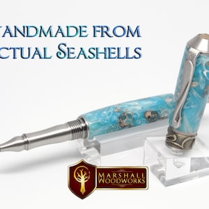 Puede incluir: Un bolígrafo azul y plateado con un diseño de conchas marinas. El bolígrafo está hecho a mano con conchas marinas reales y se muestra en un soporte de acrílico transparente. El texto "HANDMADE FROM ACTUAL SEASHELLS" es visible en la imagen. El logotipo "MARSHALL WOODWORKS" también es visible en la imagen.