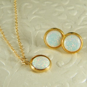 Könnte beinhalten: Ein Gold-Halsketten- und Ohrring-Set mit weißen Opal-Steinen. Die Halskette hat eine zarte Kette und einen runden Anhänger mit einem weißen Opal-Stein. Die Ohrringe sind Ohrstecker mit runden weißen Opal-Steinen.